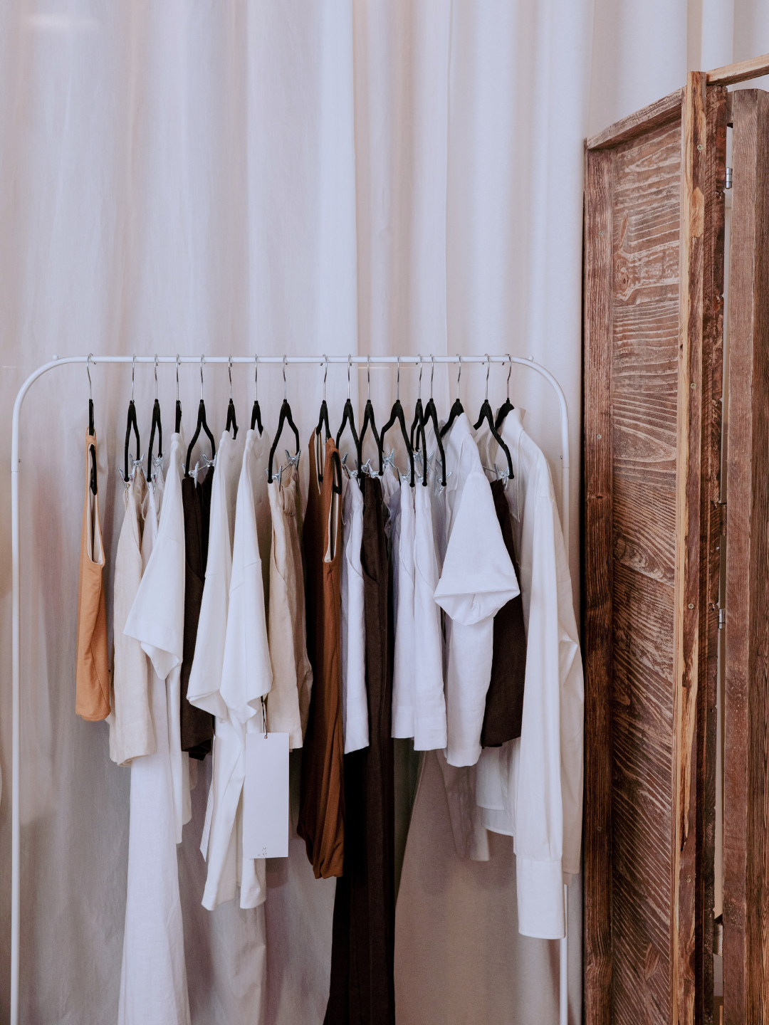 A white clothing rack holding multiple white, beige, and brown garments on black hangers, set against a soft cream curtain with a rustic wooden screen on the right. alternative wardrobe options.