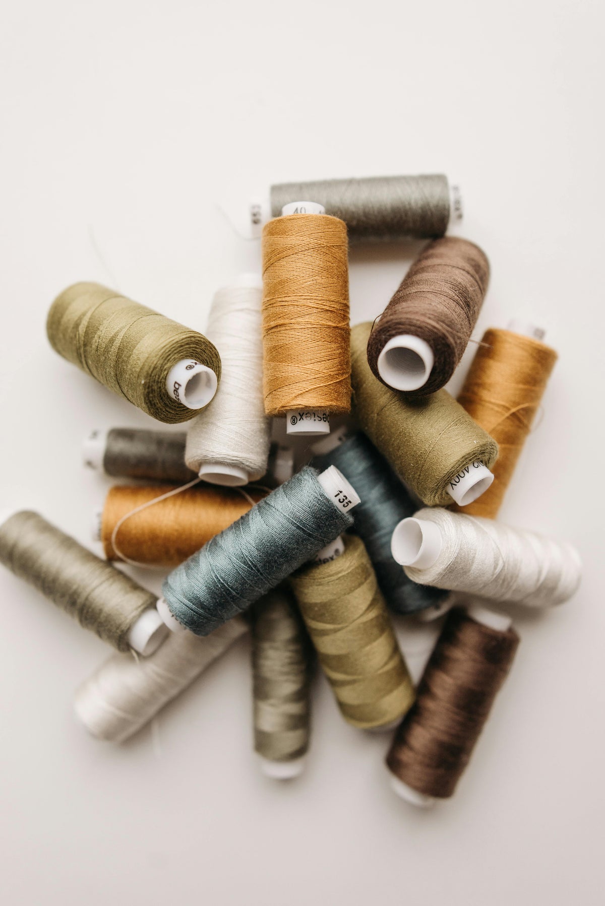 A pile of assorted sewing thread spools in muted earthy tones including olive green, dusty blue, tan, brown, cream, and gray, arranged on a light off-white background.